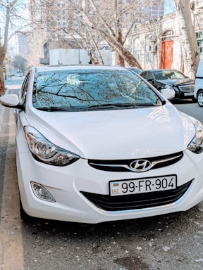 A white Hyundai car is parked on the side of a street, with bare trees and buildings in the background. The license plate reads 'AZ 99-FR-904'. Reflections of the trees are visible on the car's windshield.