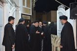 A group of men dressed in black garments gather in an outdoor setting with white gates and a partially visible building. Some men wear black head coverings, and they are engaged in conversation. Empty chairs and a speaker can be seen in the background.
