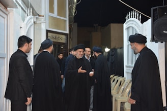 A group of men dressed in black garments gather in an outdoor setting with white gates and a partially visible building. Some men wear black head coverings, and they are engaged in conversation. Empty chairs and a speaker can be seen in the background.