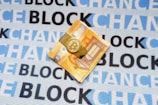 A folded euro note is secured with a Bitcoin-themed money clip, resting on a fabric background. The fabric is patterned with the repeated words 'BLOCKCHAIN' in alternating shades of blue and black.
