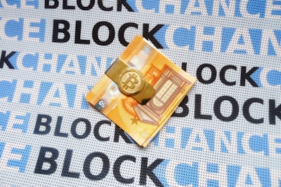 A folded euro note is secured with a Bitcoin-themed money clip, resting on a fabric background. The fabric is patterned with the repeated words 'BLOCKCHAIN' in alternating shades of blue and black.