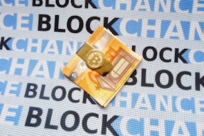 A folded euro note is secured with a Bitcoin-themed money clip, resting on a fabric background. The fabric is patterned with the repeated words 'BLOCKCHAIN' in alternating shades of blue and black.