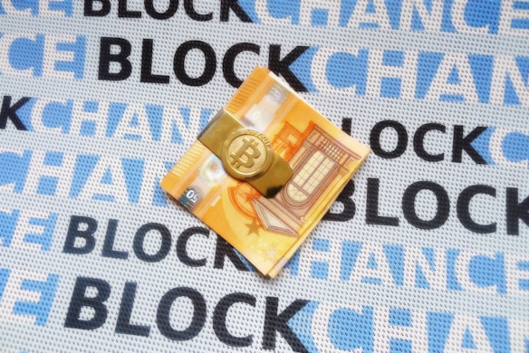 A folded euro note is secured with a Bitcoin-themed money clip, resting on a fabric background. The fabric is patterned with the repeated words 'BLOCKCHAIN' in alternating shades of blue and black.