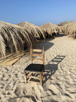 A rustic chair with a black seat is placed on a sandy beach. Surrounding the chair, there are several straw sunshades casting shadows on the ground. The clear blue sky suggests a bright and sunny day.