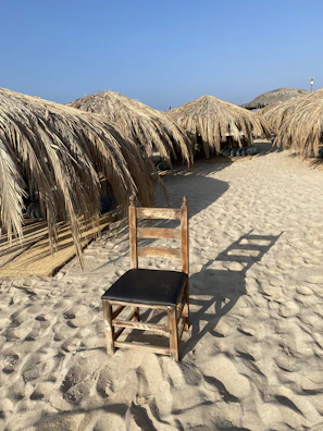A rustic chair with a black seat is placed on a sandy beach. Surrounding the chair, there are several straw sunshades casting shadows on the ground. The clear blue sky suggests a bright and sunny day.