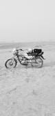 A motorcycle is parked on a sandy beach or desert area. The landscape is flat and open, with a calm and clear sky. The motorbike is positioned to the left side, and it has a helmet placed on the seat. The area is vast and isolated, contributing to a sense of solitude.