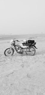 A motorcycle is parked on a sandy beach or desert area. The landscape is flat and open, with a calm and clear sky. The motorbike is positioned to the left side, and it has a helmet placed on the seat. The area is vast and isolated, contributing to a sense of solitude.