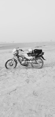 A motorcycle is parked on a sandy beach or desert area. The landscape is flat and open, with a calm and clear sky. The motorbike is positioned to the left side, and it has a helmet placed on the seat. The area is vast and isolated, contributing to a sense of solitude.