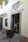A brown alpaca stands on a cobblestone street, peering into a bakery shop. The shop, named 'El Buen Pastor', has its name and details written on the wall beside the door. The bakery's display case is visible through the open door, filled with pastries.