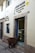 A brown alpaca stands on a cobblestone street, peering into a bakery shop. The shop, named 'El Buen Pastor', has its name and details written on the wall beside the door. The bakery's display case is visible through the open door, filled with pastries.