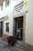 A brown alpaca stands on a cobblestone street, peering into a bakery shop. The shop, named 'El Buen Pastor', has its name and details written on the wall beside the door. The bakery's display case is visible through the open door, filled with pastries.