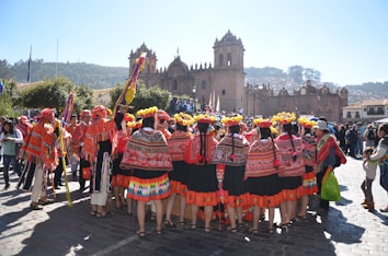 A large group of people in vibrant, traditional Andean clothing are gathered in the foreground of a historic stone plaza. The clothing features colorful patterns and designs, with many individuals wearing shawls, skirts, and hats adorned with yellow pom-poms. People are engaged in a celebration or festival, with others observing the scene. In the background, an ornate, historic stone cathedral with towers and intricate architecture overlooks the scene. Trees and distant hills complete the background.