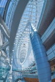 A spacious modern architectural interior with curving, sweeping lines and a series of large, overlapping arches. The structure uses a combination of glass and metal, allowing natural light to flood the space and create a bright, airy environment. The design features a large central column and intricate frameworks that contribute to the futuristic ambiance.