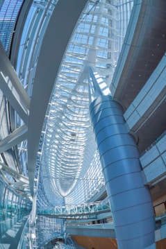 A spacious modern architectural interior with curving, sweeping lines and a series of large, overlapping arches. The structure uses a combination of glass and metal, allowing natural light to flood the space and create a bright, airy environment. The design features a large central column and intricate frameworks that contribute to the futuristic ambiance.