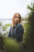 A young person with long hair stands in a natural setting, surrounded by greenery and soft-focus foliage. They are wearing a denim jacket and have a serene expression, with their eyes closed as they tilt their head slightly downward. The background features a body of water and verdant plants, creating a peaceful and tranquil scene.