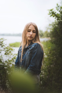 A young person with long hair stands in a natural setting, surrounded by greenery and soft-focus foliage. They are wearing a denim jacket and have a serene expression, with their eyes closed as they tilt their head slightly downward. The background features a body of water and verdant plants, creating a peaceful and tranquil scene.