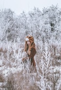 A person wearing a furry hat resembling an animal is standing in a snow-covered field. The surrounding trees are coated in frost, creating a wintery, serene landscape.