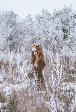A person wearing a furry hat resembling an animal is standing in a snow-covered field. The surrounding trees are coated in frost, creating a wintery, serene landscape.