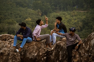 Four young men are sitting on large rocks in a natural forested area. They are engaged in conversation, and one of them is animatedly gesturing with his hand. The setting suggests they might be enjoying a casual outing or adventure.