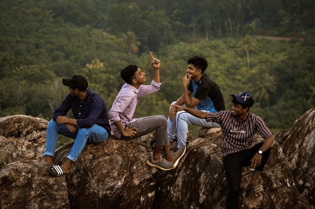 Four young men are sitting on large rocks in a natural forested area. They are engaged in conversation, and one of them is animatedly gesturing with his hand. The setting suggests they might be enjoying a casual outing or adventure.