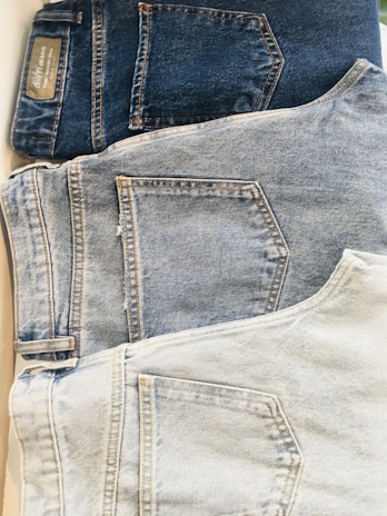 Three pairs of jeans are laid out in a row, each displaying the back pocket. The jeans range in color from dark blue at the top to light blue at the bottom. The different shades indicate varying levels of wash and wear, with stitching details visible around the pockets.