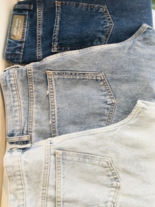 Three pairs of jeans are laid out in a row, each displaying the back pocket. The jeans range in color from dark blue at the top to light blue at the bottom. The different shades indicate varying levels of wash and wear, with stitching details visible around the pockets.