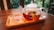 A glass teapot filled with a bright, red herbal tea sits on a wooden tray, accompanied by two empty glass teacups. The teapot contains visible flowers or herbs, lending a natural element. The background is blurred but suggests indoor greenery and a cozy, relaxed atmosphere.