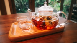 A glass teapot filled with a bright, red herbal tea sits on a wooden tray, accompanied by two empty glass teacups. The teapot contains visible flowers or herbs, lending a natural element. The background is blurred but suggests indoor greenery and a cozy, relaxed atmosphere.