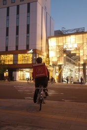 A McDonald's delivery person rides a bicycle through an urban area during twilight. The cyclist carries a branded delivery box on their back, and the streets are illuminated with streetlights and building lights. The scene features modern architecture with a mix of commercial storefronts.