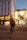 A McDonald's delivery person rides a bicycle through an urban area during twilight. The cyclist carries a branded delivery box on their back, and the streets are illuminated with streetlights and building lights. The scene features modern architecture with a mix of commercial storefronts.