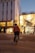 A McDonald's delivery person rides a bicycle through an urban area during twilight. The cyclist carries a branded delivery box on their back, and the streets are illuminated with streetlights and building lights. The scene features modern architecture with a mix of commercial storefronts.