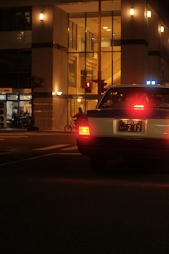 A nighttime urban scene featuring a taxi on a street with its rear lights illuminated. The taxi is positioned at an intersection in front of a modern, brightly lit building with large glass windows. A bicyclist is faintly visible, moving across the street.
