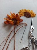 Two pieces of embroidery resembling sunflowers are displayed on a light background. One is partially completed with orange and yellow threads still attached. Beside it, there is a pair of ornate scissors and a needle.