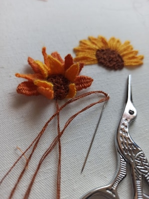 Two pieces of embroidery resembling sunflowers are displayed on a light background. One is partially completed with orange and yellow threads still attached. Beside it, there is a pair of ornate scissors and a needle.