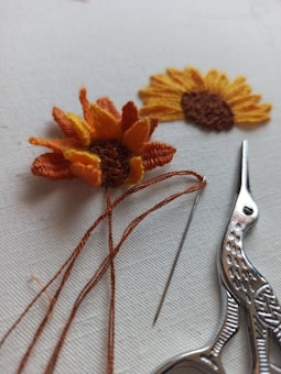 Two pieces of embroidery resembling sunflowers are displayed on a light background. One is partially completed with orange and yellow threads still attached. Beside it, there is a pair of ornate scissors and a needle.