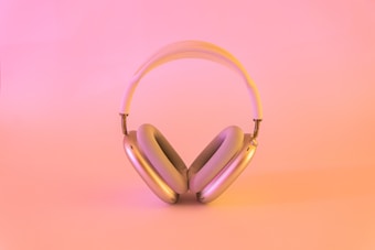 A pair of over-ear headphones with a sleek and modern design is positioned centrally on a soft, pinkish-orange background. The headphones feature a metallic finish with plush ear cushions and a padded headband.