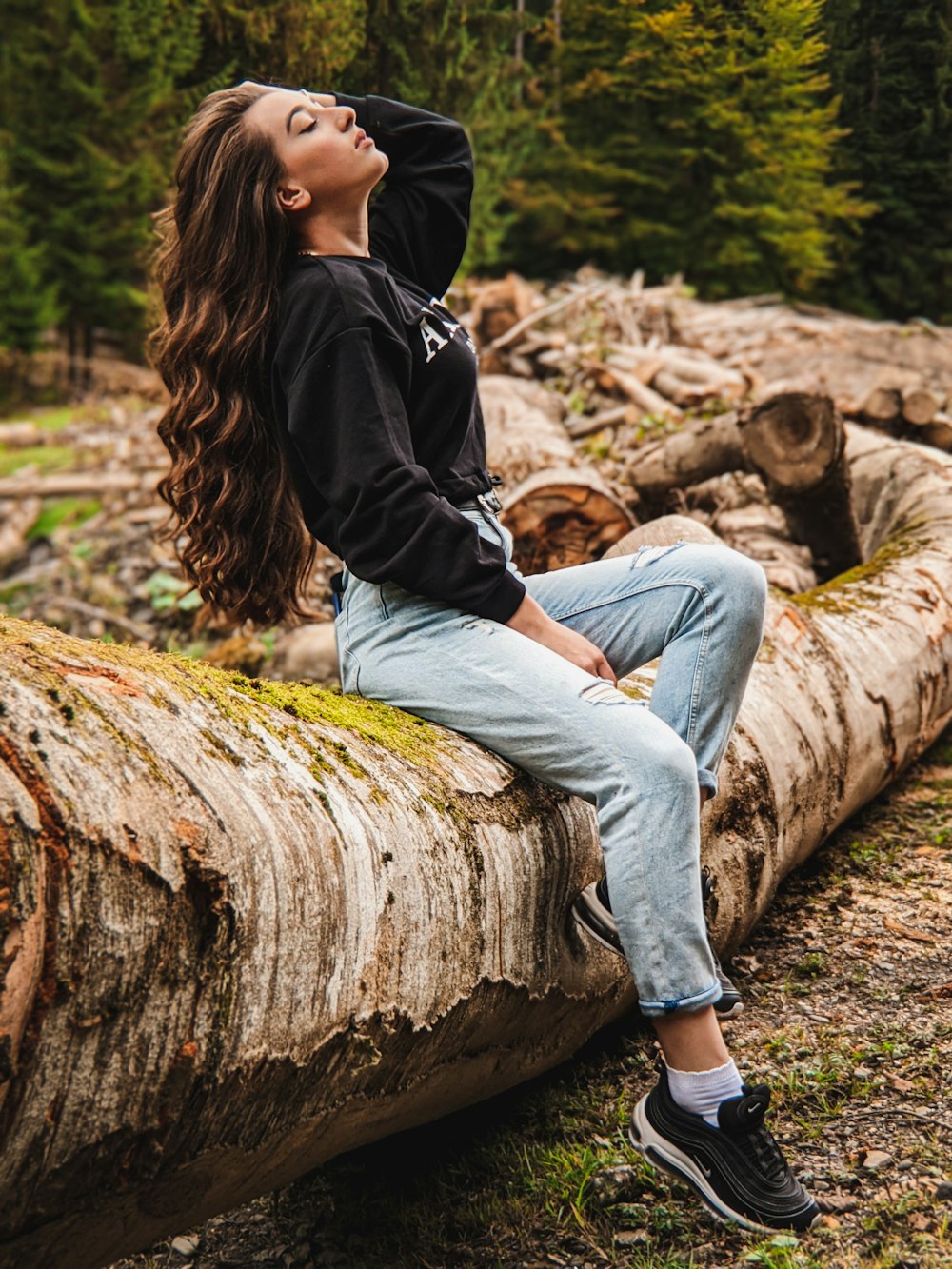 woman in black jacket and blue denim jeans sitting on brown wood log