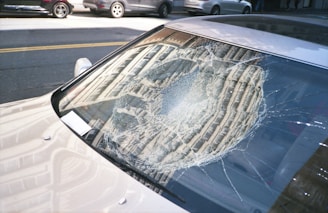 A car windshield with a large, circular crack in the center, showing spider-web patterns in the glass. The car is parked on a street with other vehicles visible in the background. Sunlight reflects off the cracked glass and the car's body.