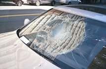 A car windshield with a large, circular crack in the center, showing spider-web patterns in the glass. The car is parked on a street with other vehicles visible in the background. Sunlight reflects off the cracked glass and the car's body.