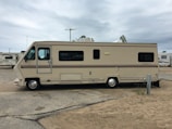 A large beige and brown motorhome is parked on a sandy area within a campground. It has a classic rectangular shape with several windows along its side. The surroundings include other RVs and motorhomes, and the sky is overcast.