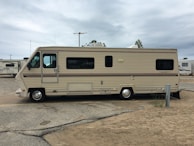 A large beige and brown motorhome is parked on a sandy area within a campground. It has a classic rectangular shape with several windows along its side. The surroundings include other RVs and motorhomes, and the sky is overcast.