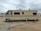 A large beige and brown motorhome is parked on a sandy area within a campground. It has a classic rectangular shape with several windows along its side. The surroundings include other RVs and motorhomes, and the sky is overcast.