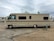 A large beige and brown motorhome is parked on a sandy area within a campground. It has a classic rectangular shape with several windows along its side. The surroundings include other RVs and motorhomes, and the sky is overcast.