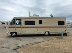A large beige and brown motorhome is parked on a sandy area within a campground. It has a classic rectangular shape with several windows along its side. The surroundings include other RVs and motorhomes, and the sky is overcast.