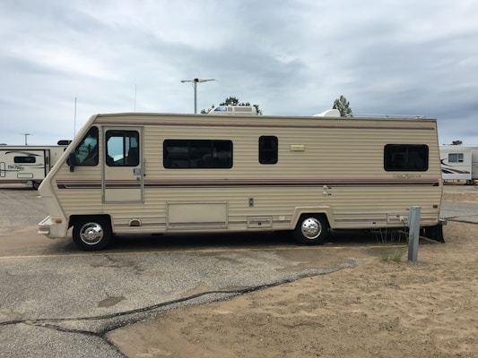 A large beige and brown motorhome is parked on a sandy area within a campground. It has a classic rectangular shape with several windows along its side. The surroundings include other RVs and motorhomes, and the sky is overcast.