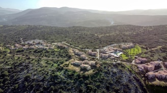 A scenic aerial view of a cluster of residential buildings nestled in a hilly landscape covered with green vegetation. The buildings are surrounded by winding roads, trees, and patches of grass, with mountains visible in the background under a hazy sky.