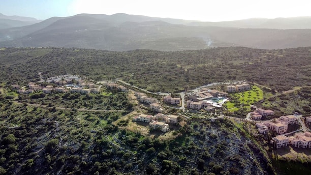 A scenic aerial view of a cluster of residential buildings nestled in a hilly landscape covered with green vegetation. The buildings are surrounded by winding roads, trees, and patches of grass, with mountains visible in the background under a hazy sky.