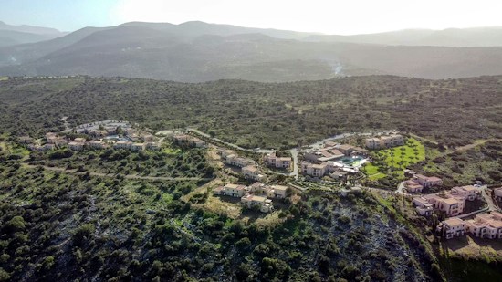 A scenic aerial view of a cluster of residential buildings nestled in a hilly landscape covered with green vegetation. The buildings are surrounded by winding roads, trees, and patches of grass, with mountains visible in the background under a hazy sky.