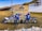 Two people wearing blue jackets and helmets stand in front of a large wooden sign that reads 'Bolognola-Ski, Parco Monti Sibillini'. They are holding hands with their backs to the camera, and each is alongside a blue and white motorcycle. The ground is grassy with patches of snow around, and there are snow-covered hills in the background.