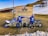 Two people wearing blue jackets and helmets stand in front of a large wooden sign that reads 'Bolognola-Ski, Parco Monti Sibillini'. They are holding hands with their backs to the camera, and each is alongside a blue and white motorcycle. The ground is grassy with patches of snow around, and there are snow-covered hills in the background.